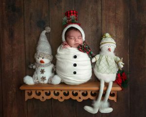 Swaddled newborn dressed as snowman posed on wooden shelf flanked by two holiday snowman figurines.