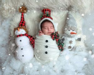Sleeping newborn wrapped in white knit swaddle posed between two snowman dolls with festive holiday decorations.