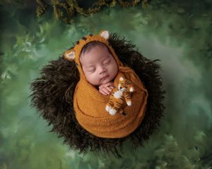 Newborn baby wrapped in orange knit swaddle with tiger bonnet, sleeping in dark fur bowl on green background.