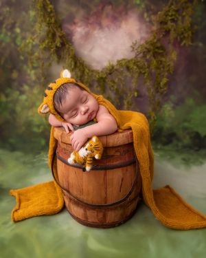 Sleeping newborn in orange lion bonnet and wrap, posed in rustic wooden barrel with tiger toy on green backdrop.