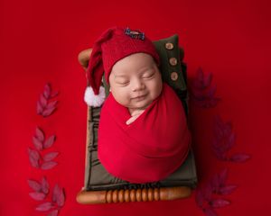Smiling newborn swaddled in red wrap with Santa hat on wooden bed.