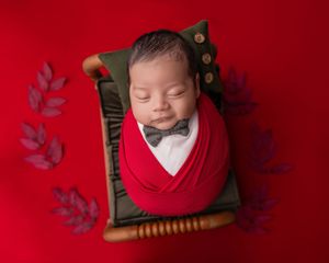 Smiling newborn baby wrapped in red swaddle wearing bow tie, sleeping in small wooden bed on red background.