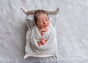Peacefully sleeping newborn swaddled in white wrap lying on a miniature white wooden bed.
