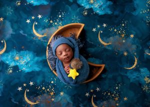 Sleeping newborn wrapped in blue on a crescent moon with starry night background.