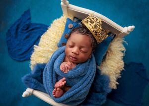 Newborn wrapped in blue swaddle wearing a gold crown on a tiny white bed.