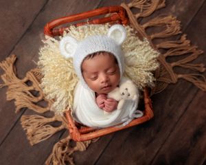 Sleeping newborn in white bear bonnet and wrap cuddling a teddy bear in a wicker basket.