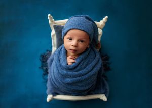 Newborn baby wrapped in blue knit blanket wearing matching hat lying in a small white wooden bed on blue backdrop.