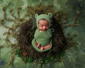 Smiling newborn baby in green frog outfit and bonnet posed in a mossy nest on green backdrop.