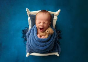 Yawning newborn baby wrapped in blue swaddle hugging a teddy bear in a tiny white bed on a teal background.