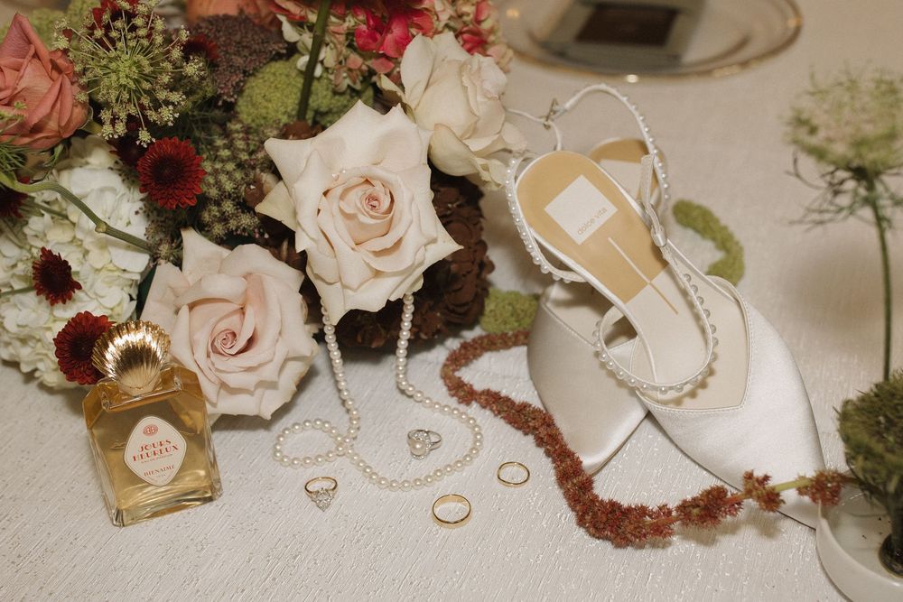 Wedding details including blush roses, rings, and white bridal shoes arranged on a light surface with delicate styling.
