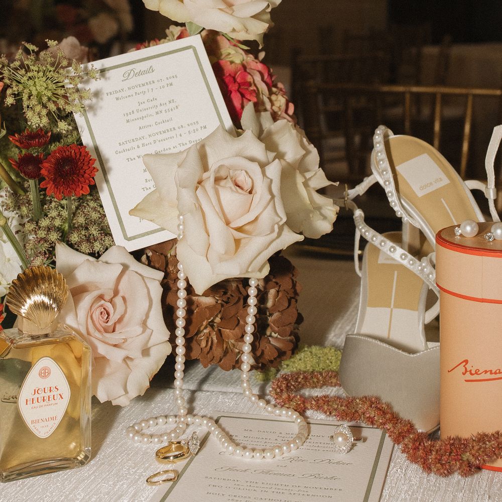 Delicate blush roses and vintage-style wedding decor arranged on a rustic wooden table with invitation cards and pearls.