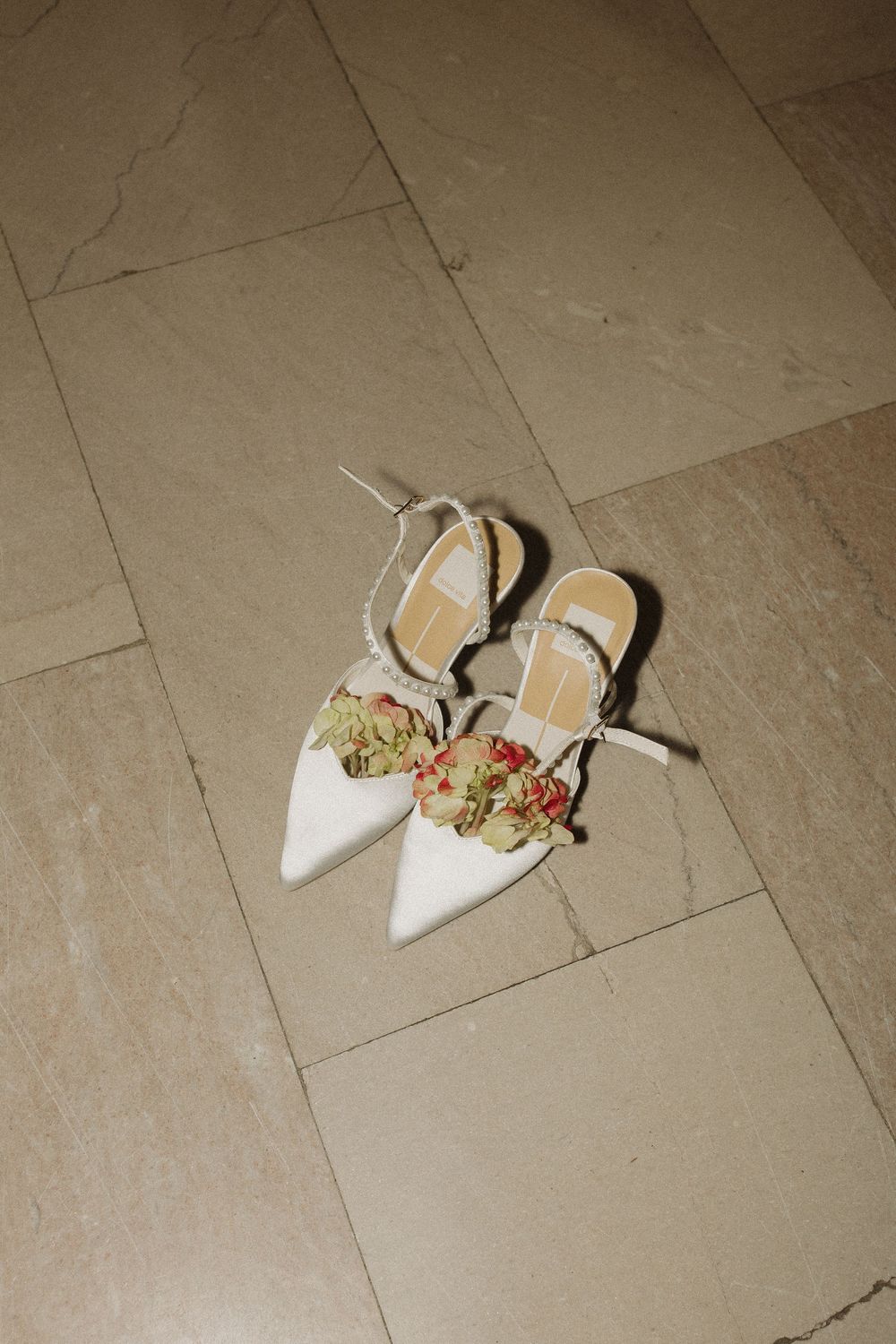 White pointed-toe slingback heels with floral decorative elements arranged on beige tile flooring.