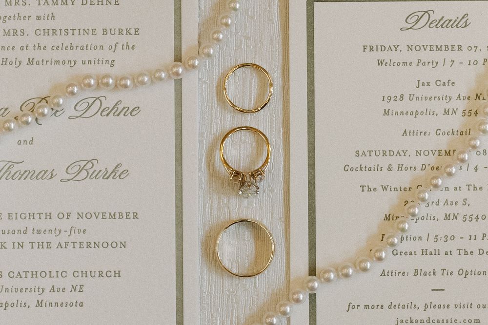 Wedding rings and engagement ring arranged on elegant ivory invitation cards with gold details and pearl accents.