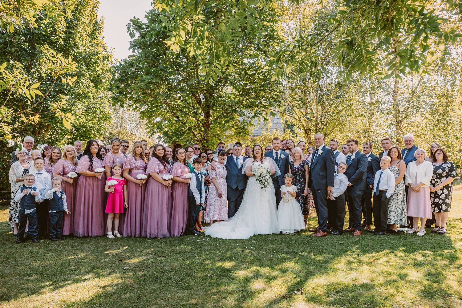 A wedding party and guests gather outdoors in a sunny garden setting for group photos.