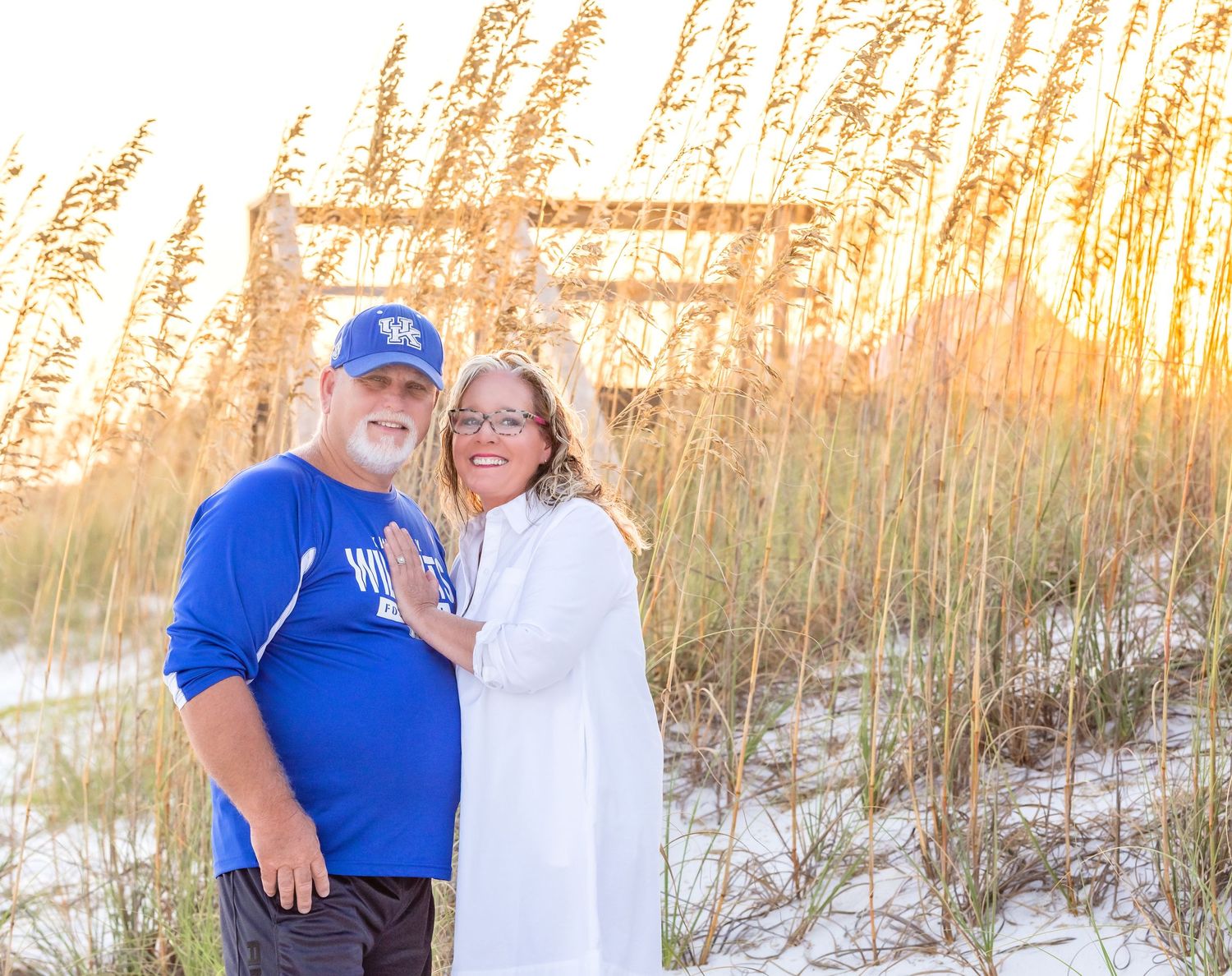 A couple stands together on a beach at sunset with tall sea oats swaying in the golden light.