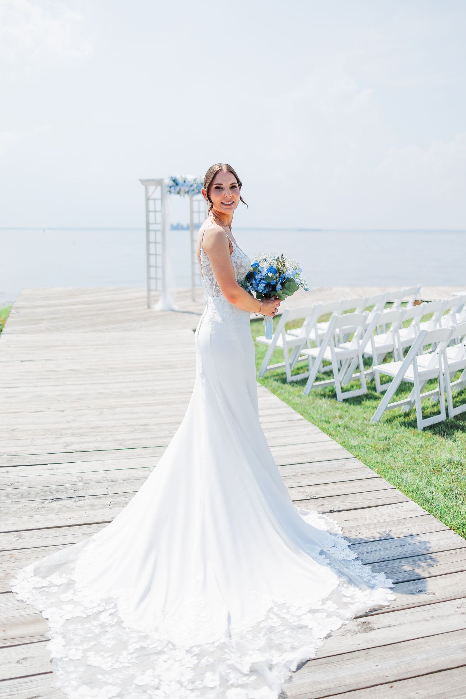 A bride in a flowing white mermaid gown holds a blue bouquet at a waterfront wedding ceremony setup with white chairs.