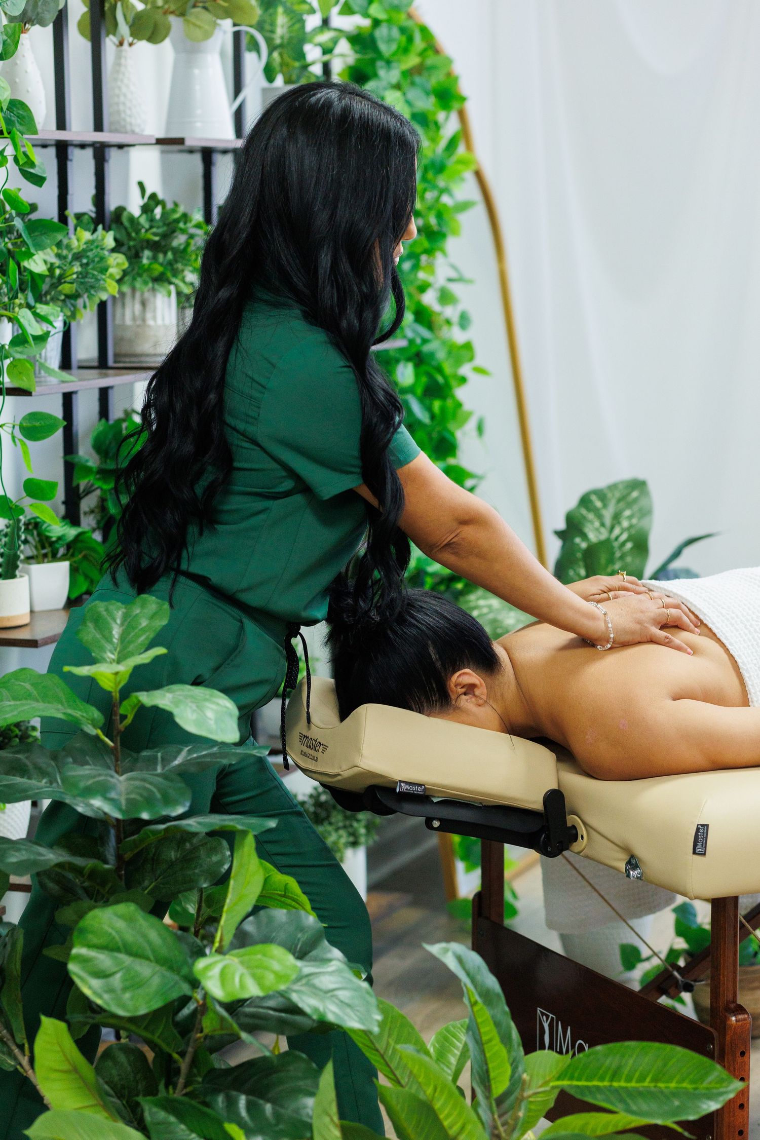 Massage therapist in green uniform performs back massage in plant-filled wellness studio.