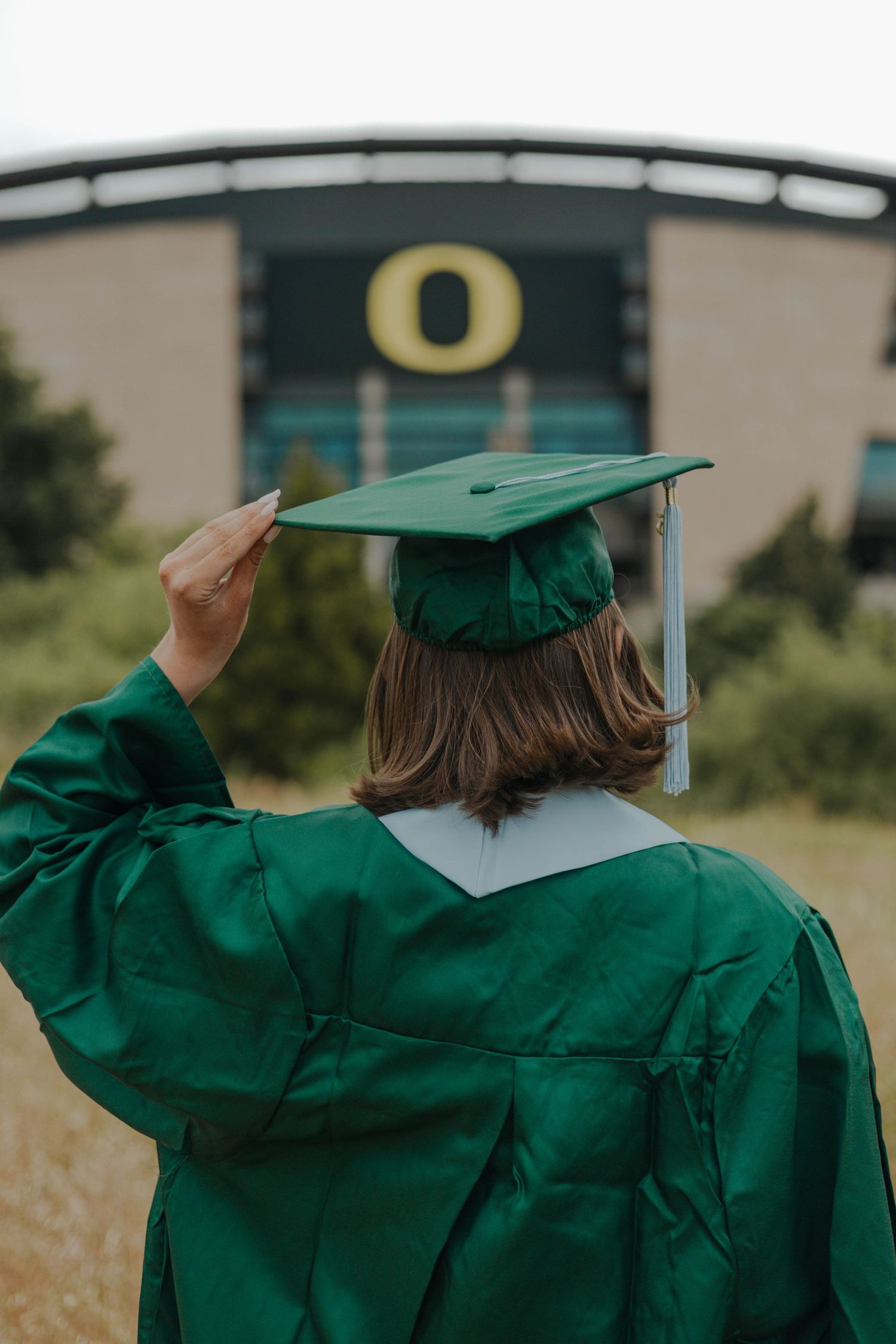 Graduate in green cap and gown looks toward University of Oregon O logo on stadium building in background.