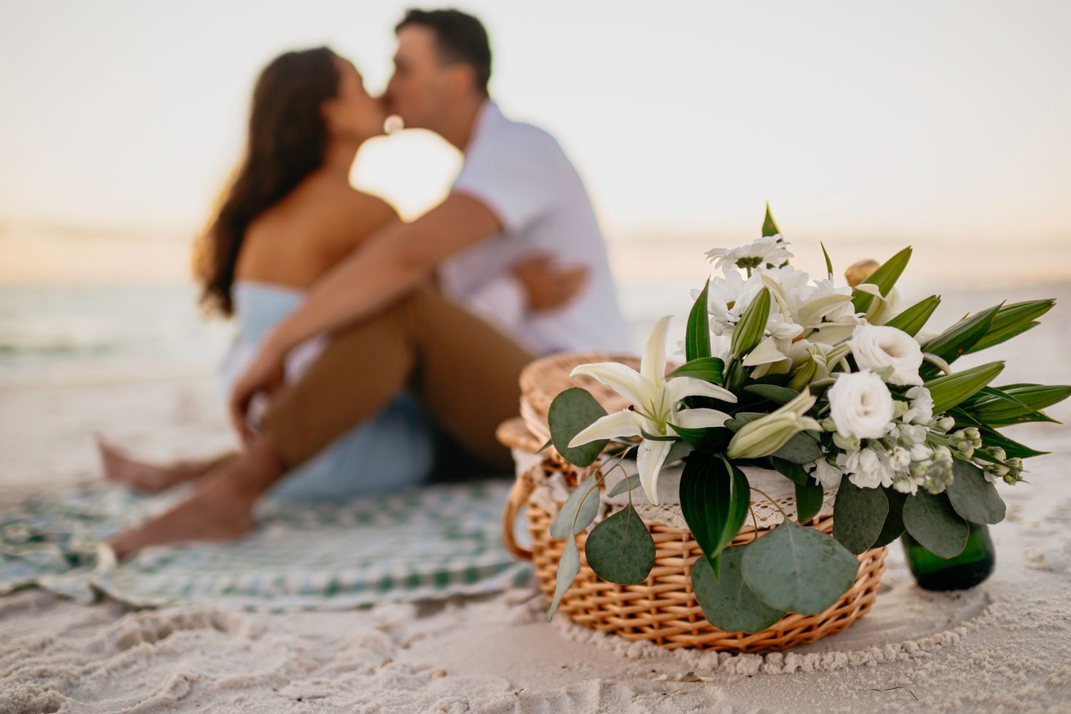Wedding rings and floral arrangements with eucalyptus leaves displayed on sandy beach at sunset.
