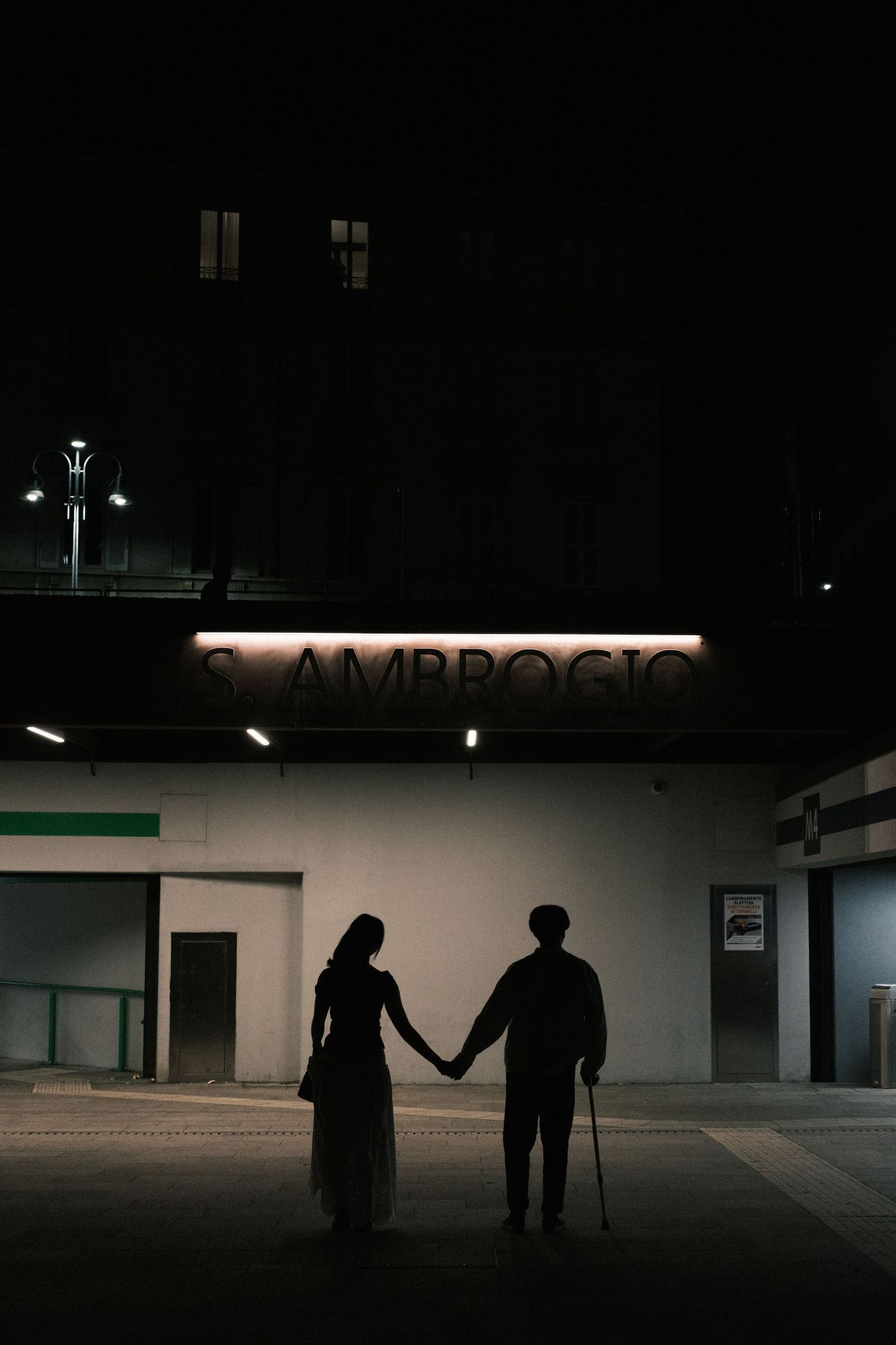 Silhouetted couple embracing under illuminated Ambrogio sign at night against dark building facade.