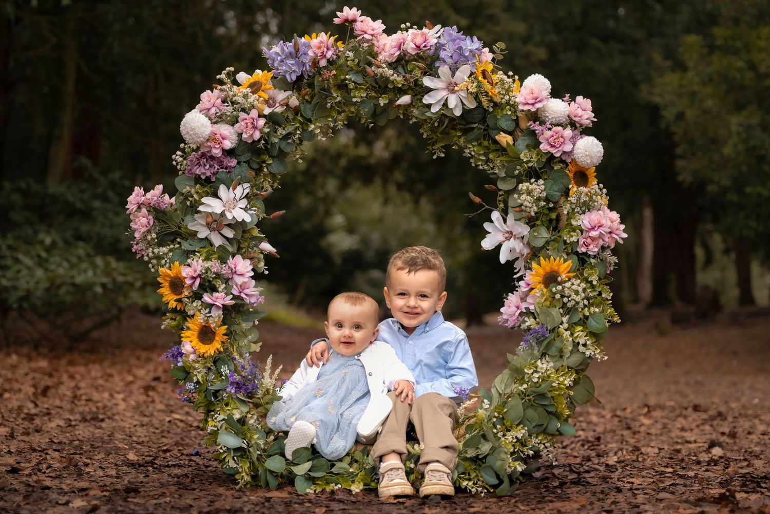 A floral arch decorated with pink and purple blooms stands in a wooded outdoor setting during a photo session.