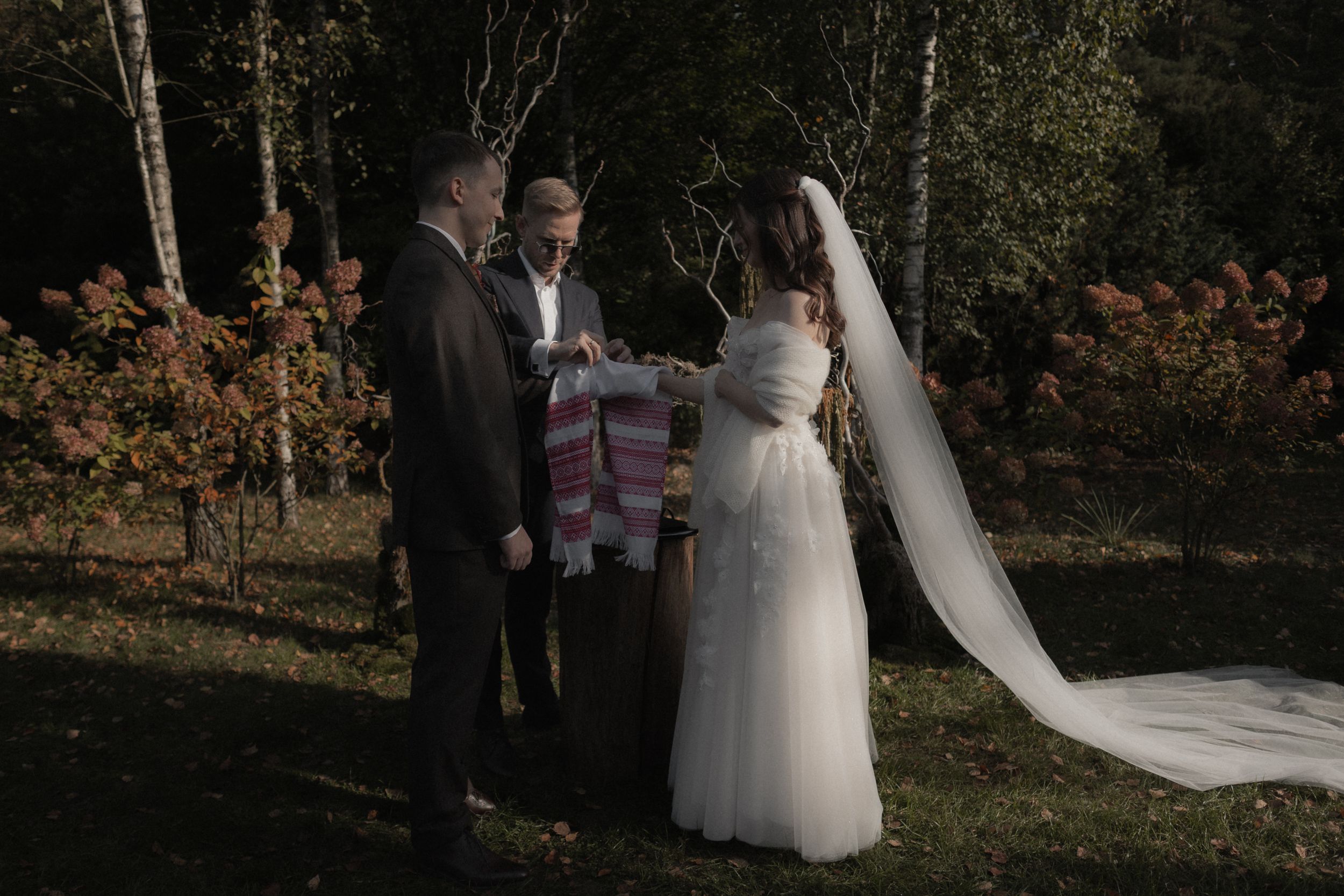 A wedding couple walks through a wooded forest setting with sunlight filtering through the trees.