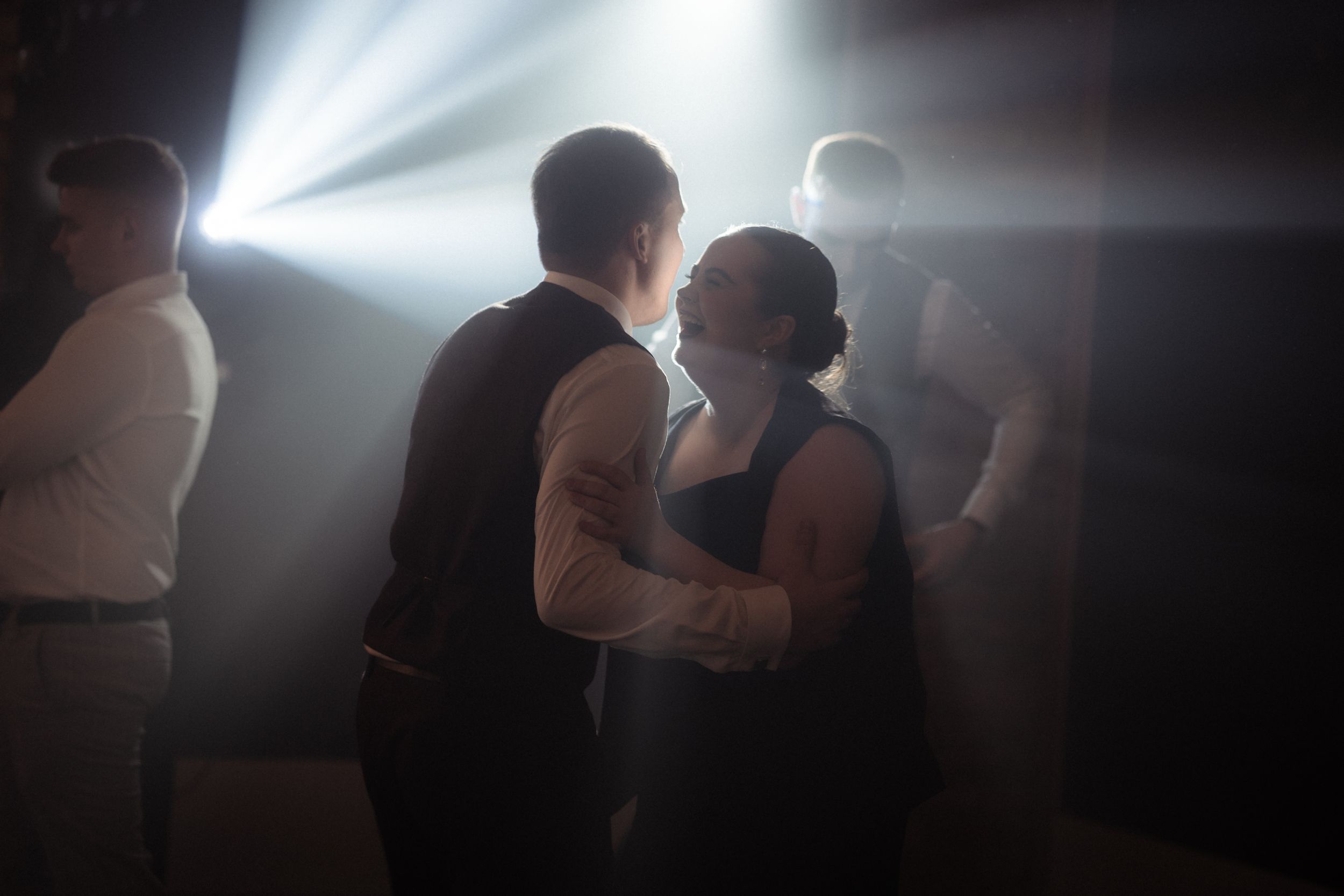Couples dance together in a dimly lit reception venue with dramatic lighting creating silhouettes.
