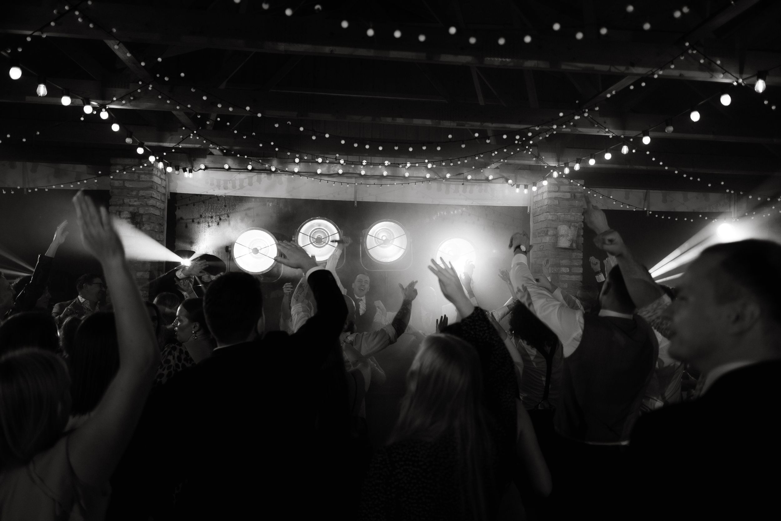 Black and white photo of a crowded dance floor at a reception with string lights overhead and bright spotlights glowing.