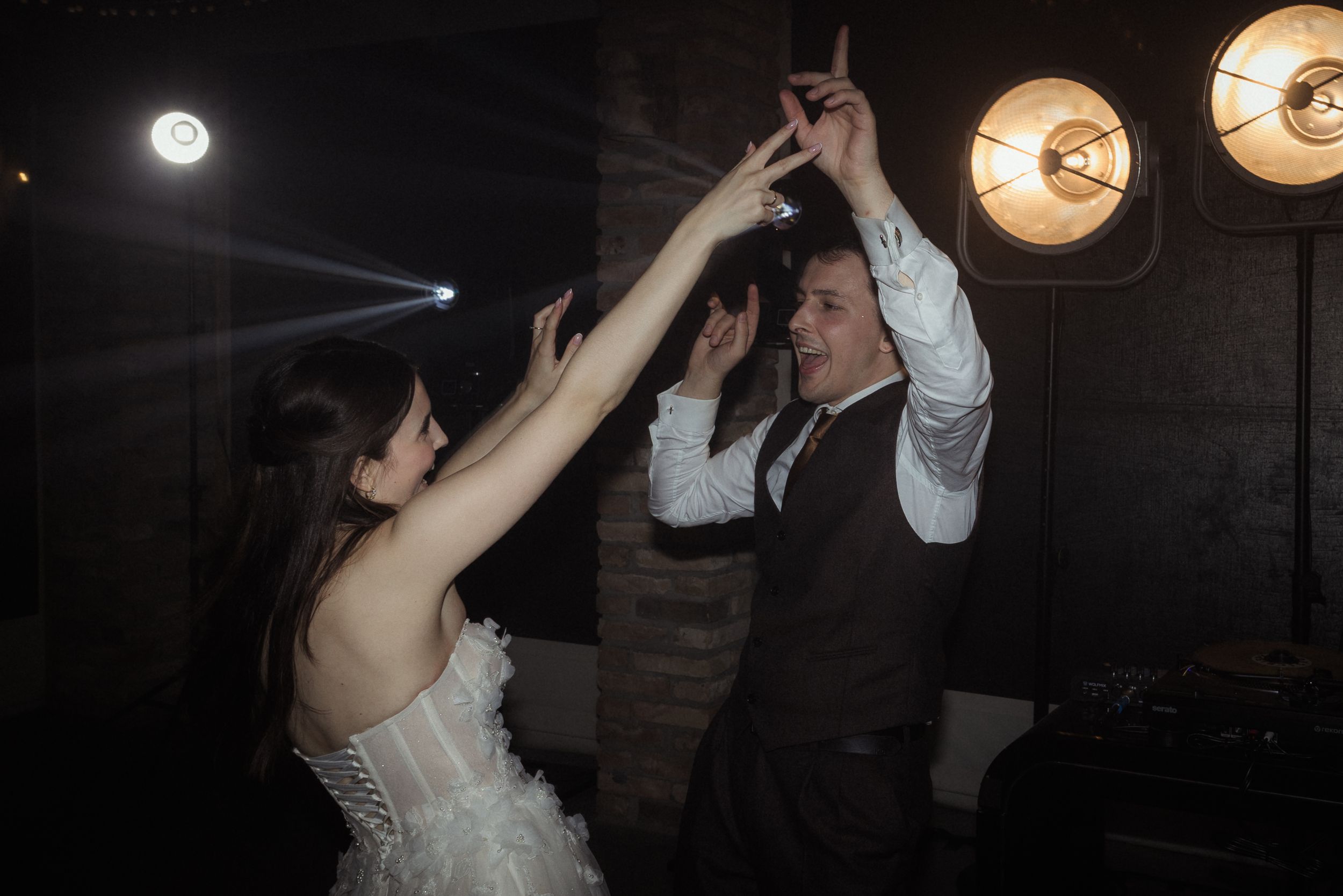People dancing and celebrating at a dimly lit wedding reception with warm glowing lights overhead.