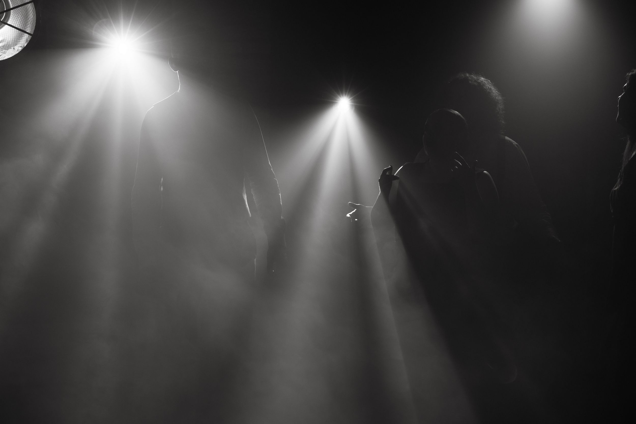 Dramatic beams of bright white stage lighting shine down through darkness creating atmospheric spotlights.