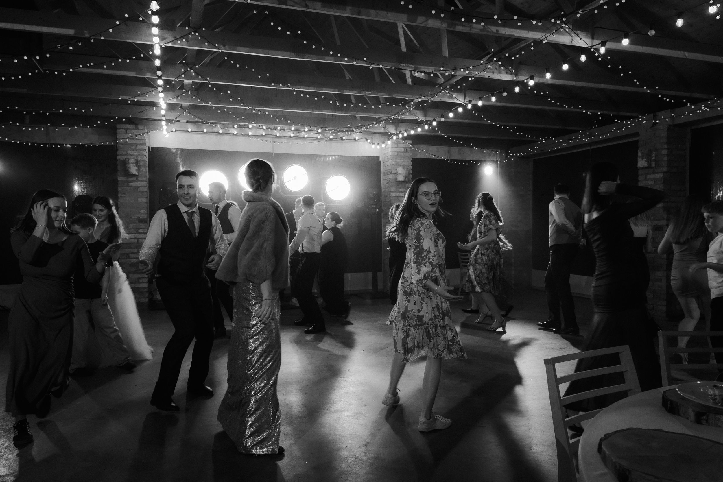 Black and white photo of people dancing at a wedding reception under string lights in a dark venue.