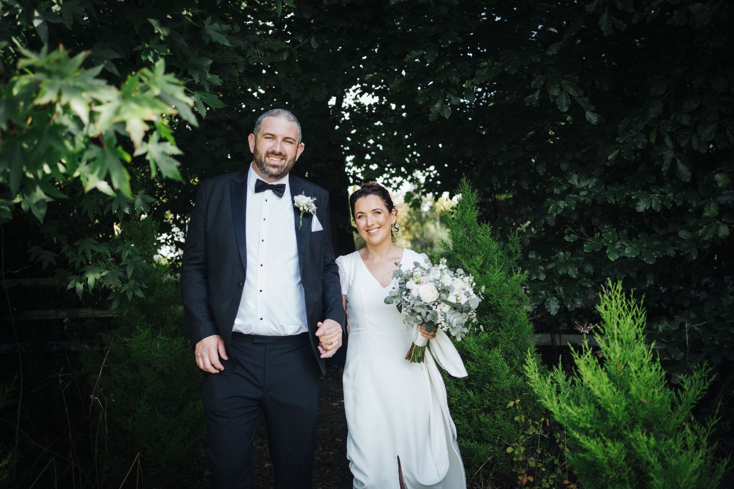 A bride and groom stand together outdoors holding hands against a lush green garden backdrop.