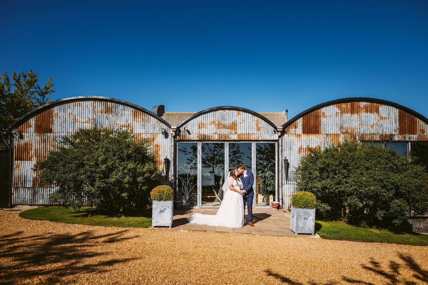 Rustic quonset hut wedding venue with arched metal buildings and gravel paths surrounded by green trees.