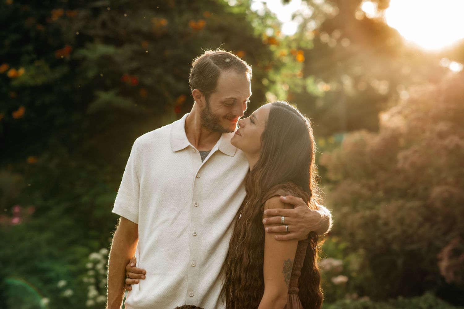 A couple embraces in a romantic sunset moment with golden sunlight filtering through trees in a natural outdoor setting.