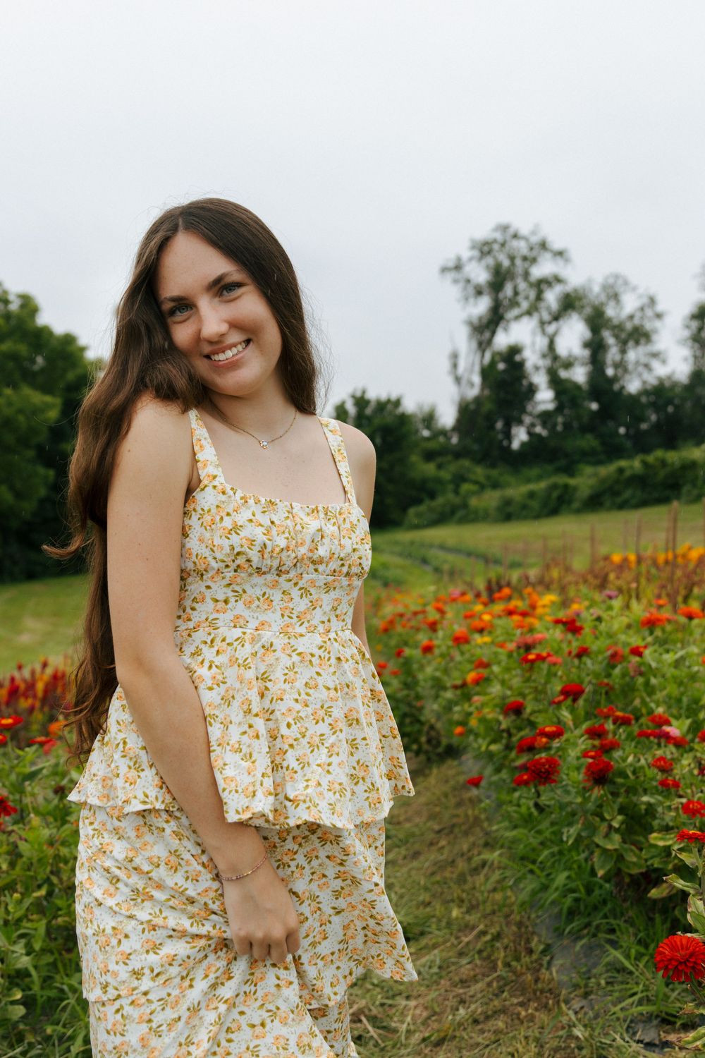 A floral sundress-clad figure poses in a vibrant garden with orange and red zinnias on a sunny summer day.