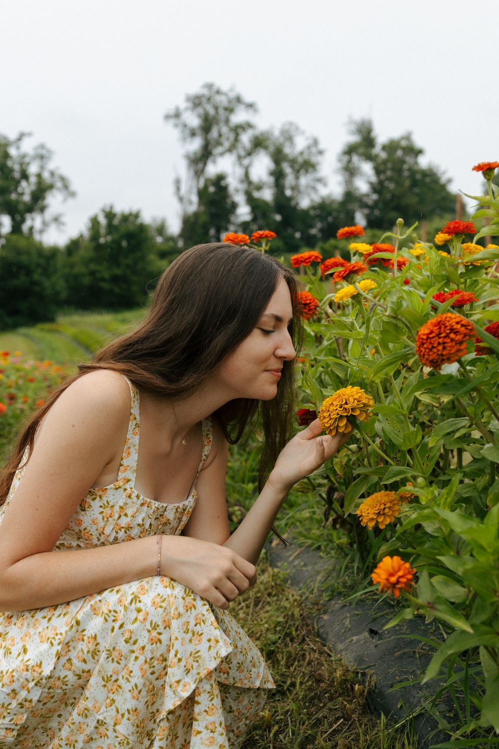 Someone in a floral dress leans in to smell bright orange zinnias growing in a garden.