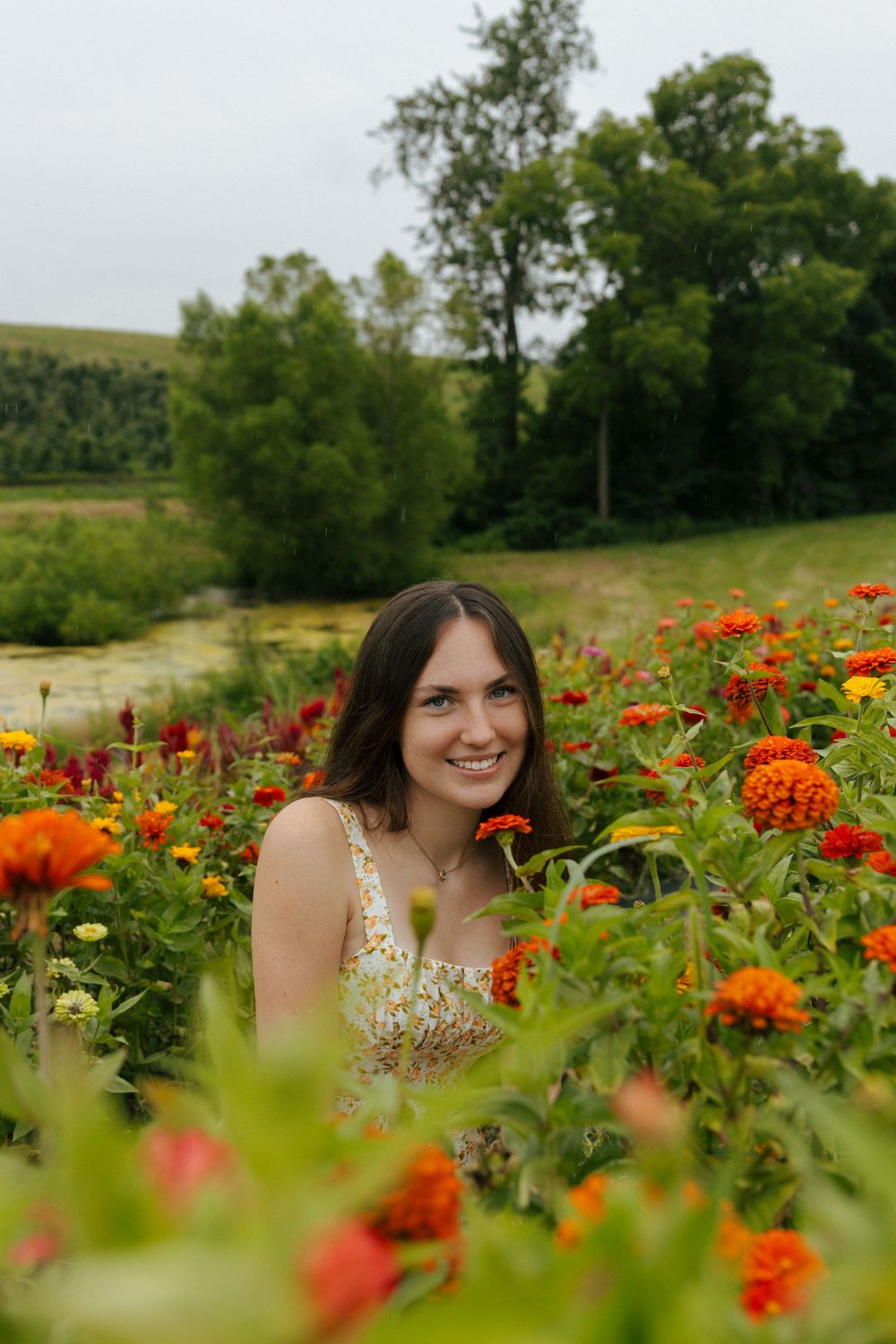 Bright orange and red zinnias surround a figure in a delicate floral sundress on a sunny day.