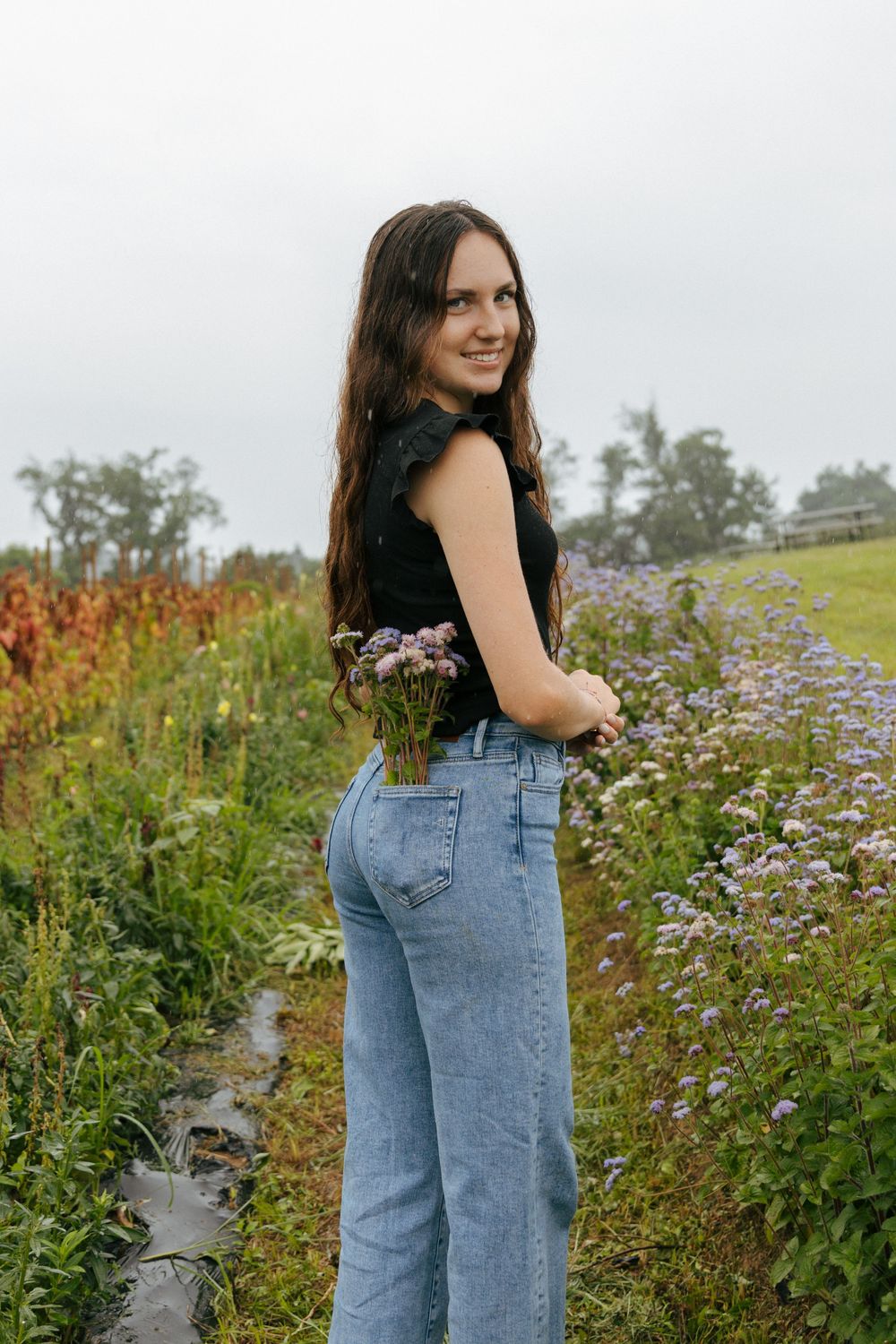 A casual outfit featuring blue jeans and dark top stands among purple wildflowers in a garden setting.