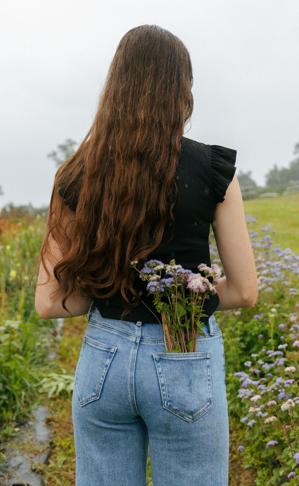 Person wearing black top and jeans poses in wildflower field with back turned to camera.