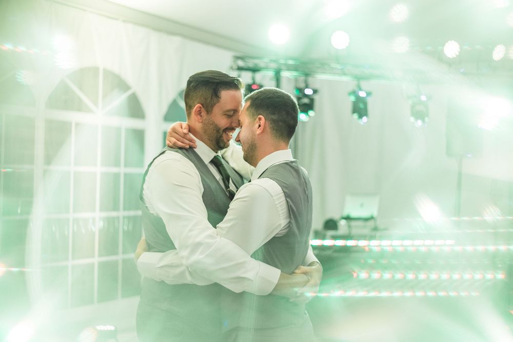 Two grooms share their first dance at a wedding reception, embracing under soft green stage lighting.