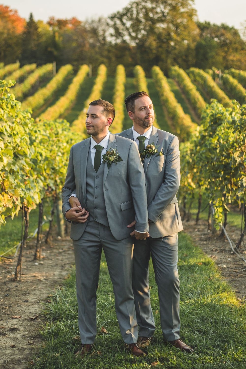 Two grooms in gray suits with boutonnieres pose in a lush vineyard on their wedding day.