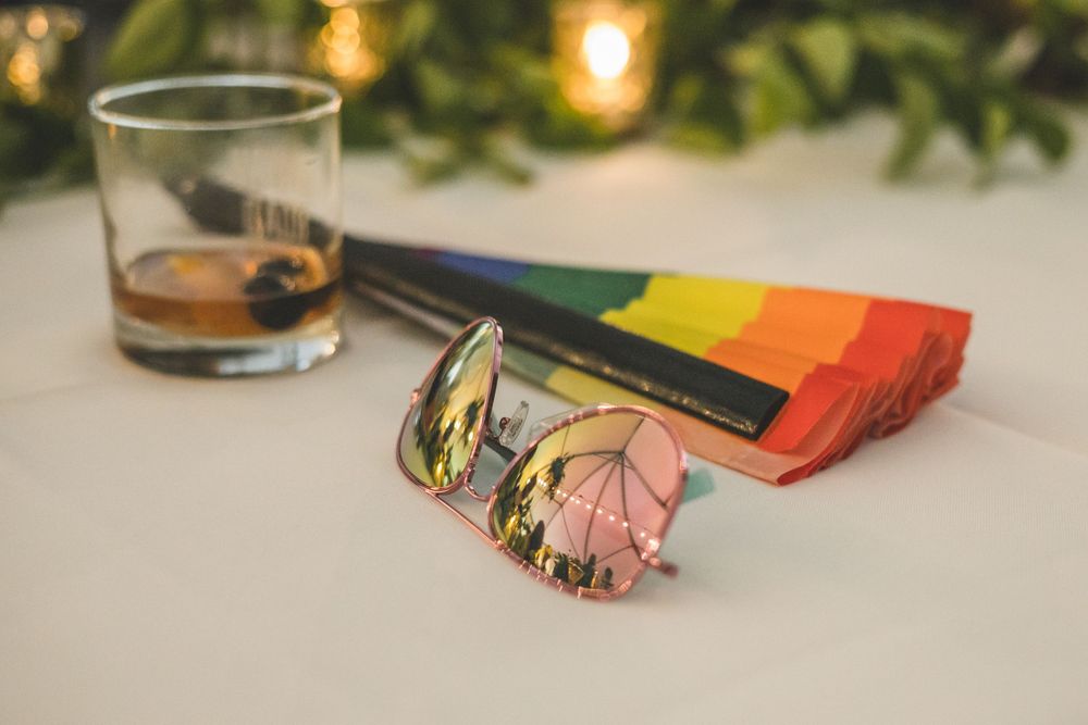 Rainbow sunglasses case and mirrored sunglasses beside a whiskey glass on a white table.