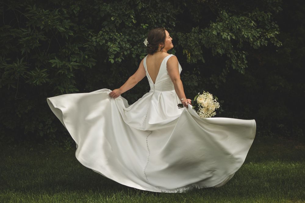 Bride in white gown twirling outdoors, holding bouquet, with flowing skirt and lush green trees in background.