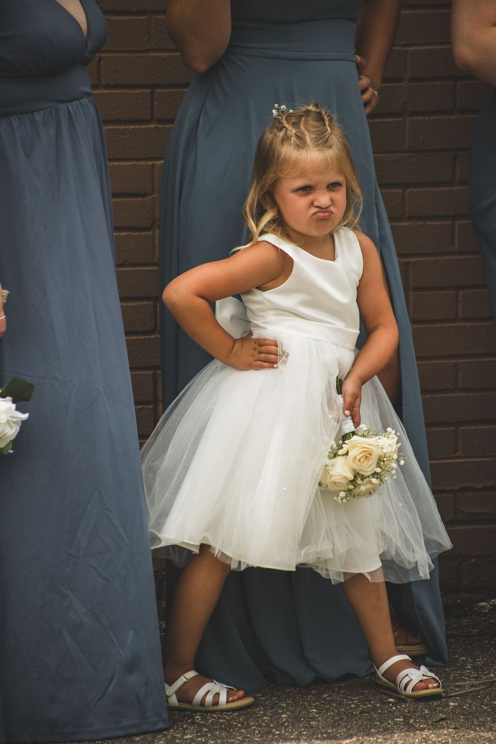 A flower girl in a white tulle dress holds a bouquet, pouting with hands on hips at a wedding ceremony.