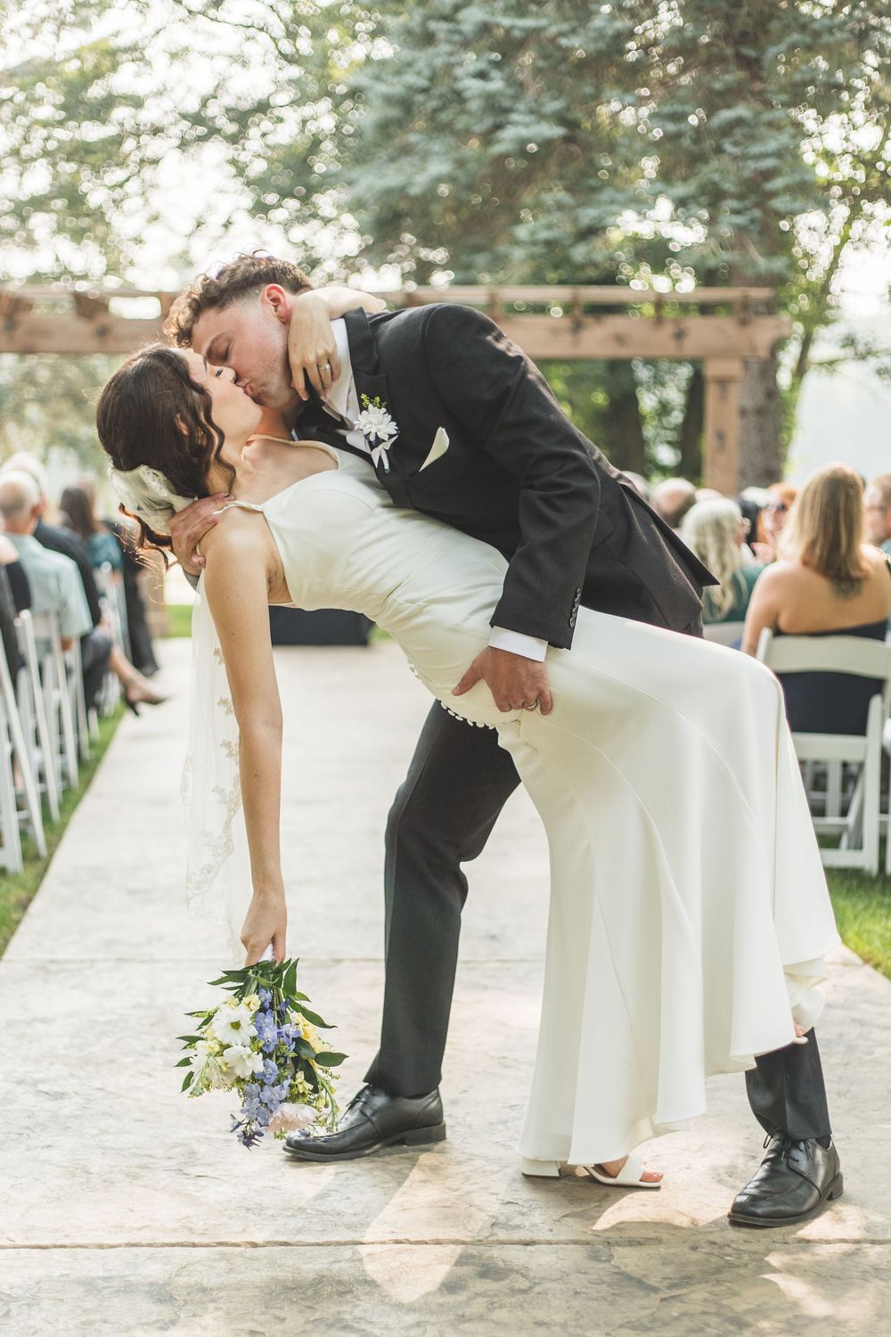 Groom dips bride for a romantic kiss at outdoor wedding ceremony while she holds a white and blue bouquet.