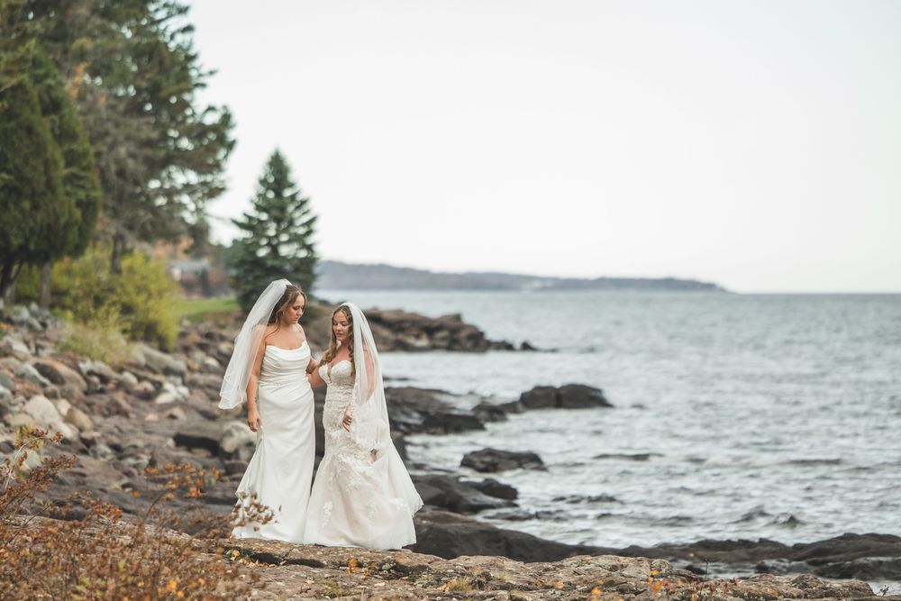 Two brides in white wedding gowns walk along a rocky shoreline with trees and calm water behind them.