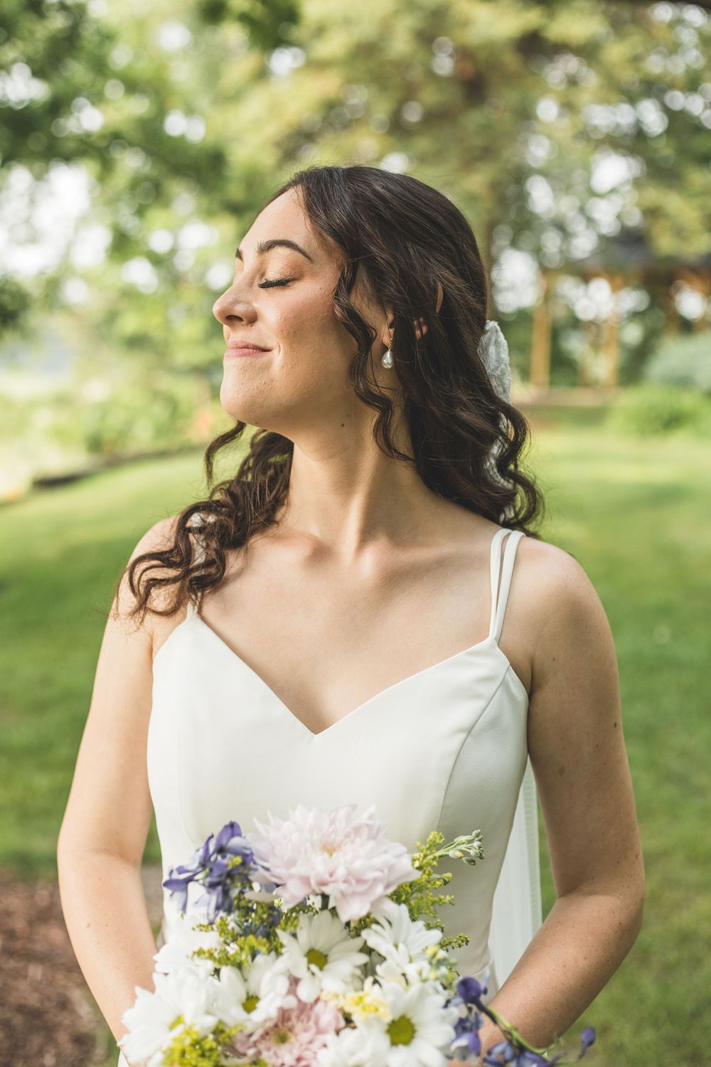 Bride in white dress holding colorful bouquet of white and purple flowers, smiling with eyes closed in lush green garden.