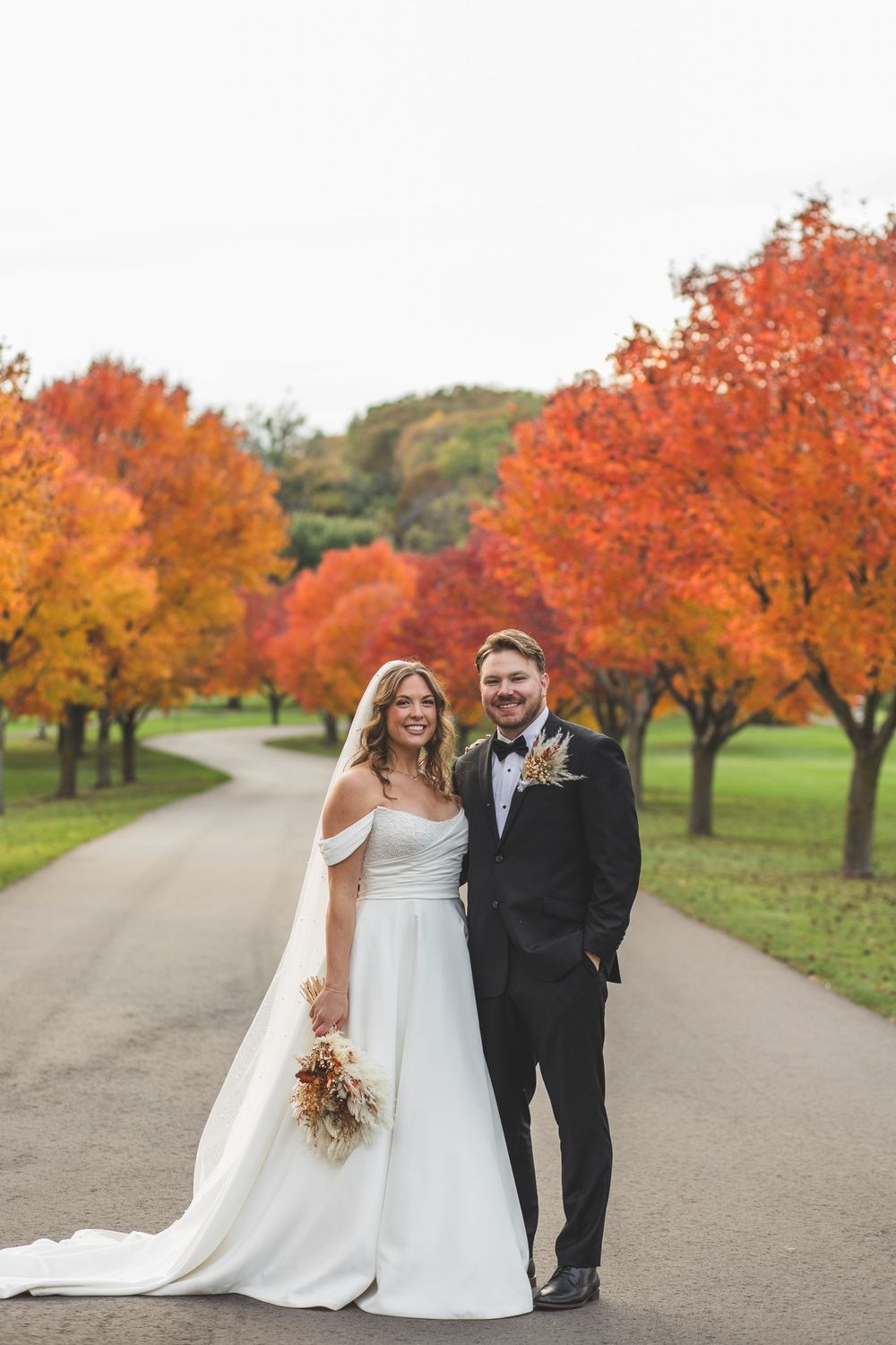 A wedding party stands on a winding path surrounded by vibrant orange and red autumn trees in a park setting.