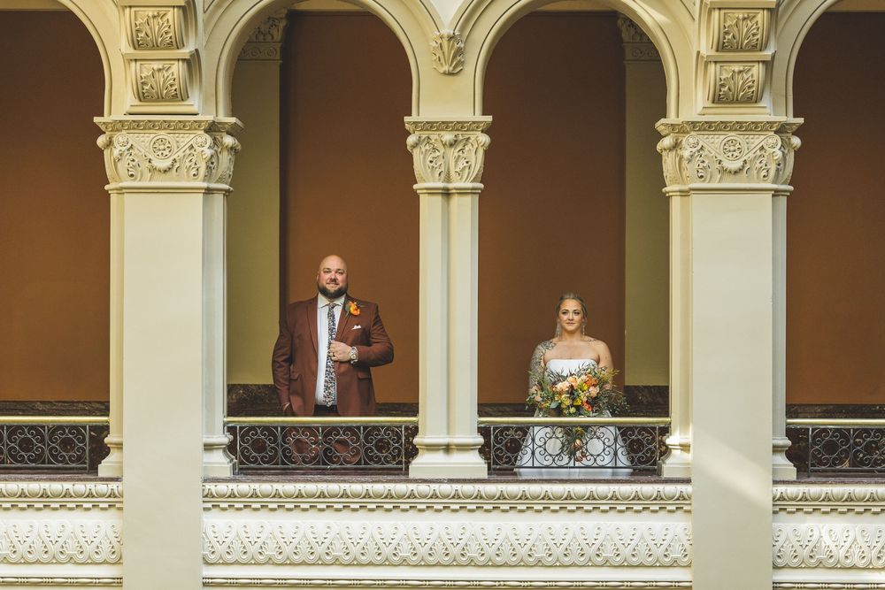 A couple stands on an elegant balcony with ornate white columns and decorative architectural details.