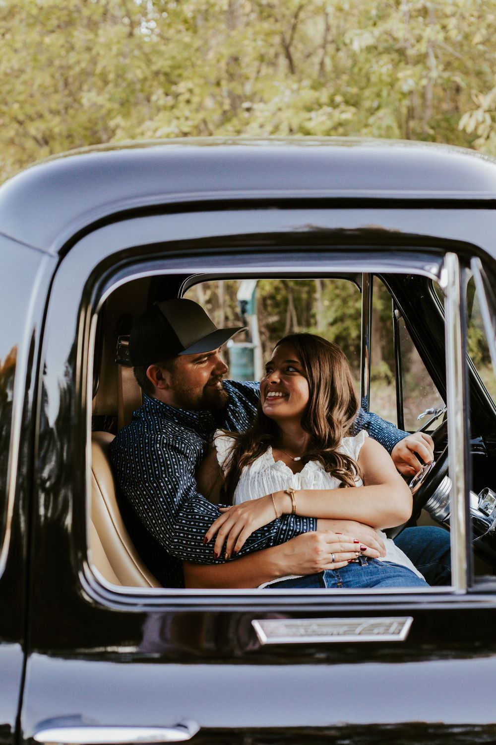 A couple shares a joyful moment together inside a vintage black car on a sunny day.